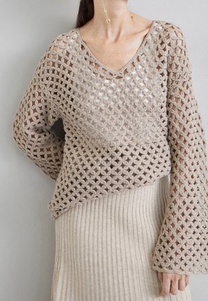 Frau trägt beige offenen Häkelpullover über weißem Top und beigem geripptem Rock, mit sichtbarem langem hängendem goldenen Ohrring.