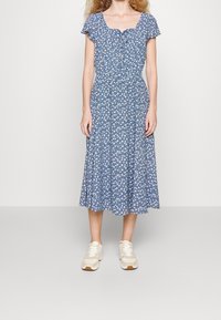 Blå blommig midiklänning med fyrkantig hals och korta ärmar, med en sammandraget midja och utställd kjol. Kombinerad med ljusa sneakers.