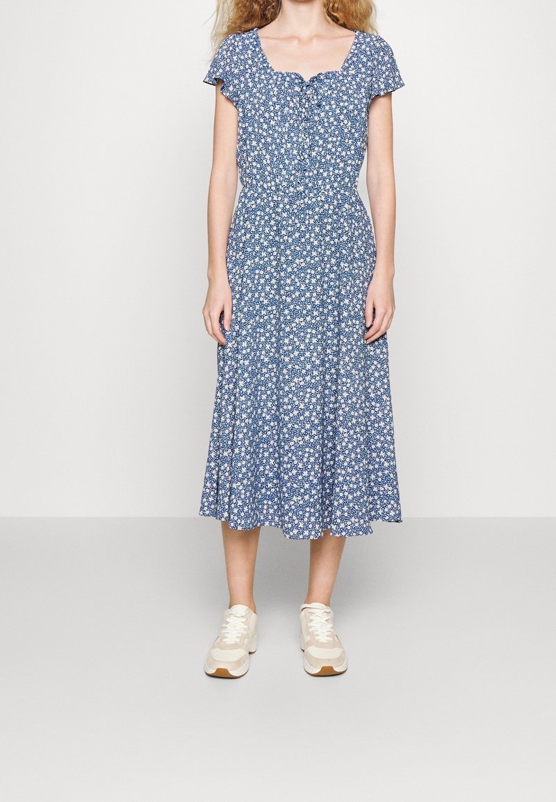 Blå blommig midiklänning med fyrkantig hals och korta ärmar, med en sammandraget midja och utställd kjol. Kombinerad med ljusa sneakers.