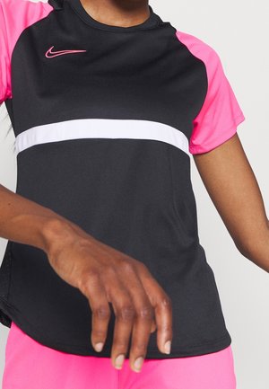 Personne portant un maillot de sport à manches courtes noir et rose avec une bande horizontale blanche et un short rose assorti, bras tendu vers l'avant.