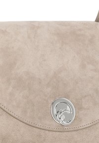 Borsa a mano in suede beige con flap curvo, caratterizzata da una chiusura circolare in argento e cuciture visibili lungo i bordi. Texture liscia.