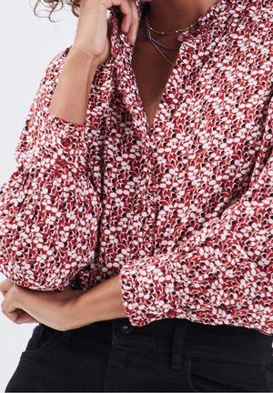 Blusa a fiori rossa e bianca con colletto alto, chiusura a bottoni e maniche a sbuffo, realizzata in tessuto leggero. Pantaloni neri visibili.