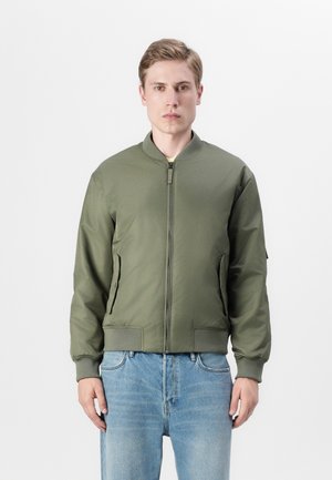 Veste bomber vert olive avec fermeture éclair sur le devant, poignets côtelés, deux poches latérales et une finition lisse et mate. Portée avec un jean en denim bleu clair.