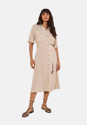 Beige Leinenhemdkleid mit kurzen Ärmeln, Knopfleiste, zwei Brusttaschen und einem Taillengürtel, ergänzt durch schwarze Sandalen mit Riemen.