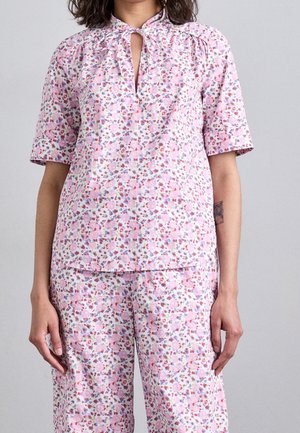 Mujer con camiseta de manga corta con estampado floral en rosa y púrpura y pantalones a juego, de pie frente a un fondo liso gris claro.
