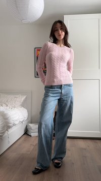 Kvinne iført en rosa kabelstrikket genser, vide blå jeans og svarte flate sko, stående i et minimalistisk soverom med hvite vegger.