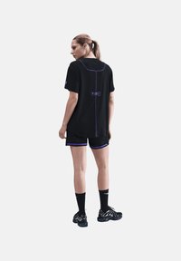 Kvinde med hestehale iført sorte atletikshorts, en matchende løs t-shirt med lilla syninger, sorte Nike-sokker og sorte sneakers med hvide detaljer.