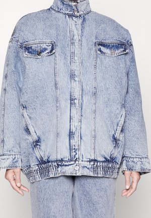 Light blue acid-wash denim jacket with front pockets, snap button closure, elastic hem, worn with matching jeans, hands relaxed at sides.