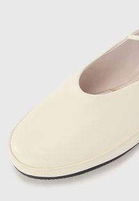 Chaussure en cuir crème avec un bout rond, au design minimaliste et une semelle en caoutchouc noire contrastante. Texture lisse avec des détails de couture subtils.