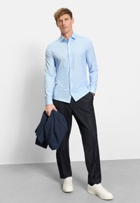 Man wearing light blue striped shirt and black trousers holding a folded navy jacket, standing against a plain light background.