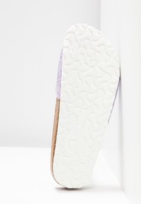 Semelle texturée blanche d'une sandale avec une semelle intermédiaire en liège et une partie supérieure lilas présentant un motif subtil, posée en angle contre un mur d'angle blanc.
