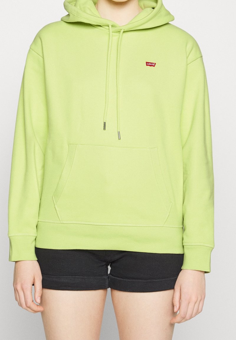 Personne portant un sweat à capuche Levi’s vert clair avec poche avant et un short noir, debout devant un fond clair uni.