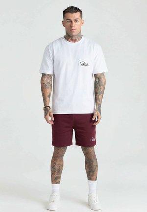 Muž s rozsiahlymi tetovaniami, oblečený v bieliom tričku a bordových kraťasoch, stojí tvárou vpred na jednoduchom svetlom pozadí.