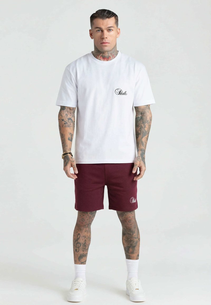 Ein Mann mit umfangreichen Tattoos, der ein weißes T-Shirt und bordeauxrote Shorts trägt, steht mit dem Gesicht nach vorne auf einem schlichten hellen Hintergrund.