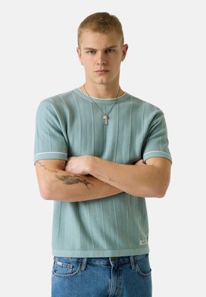 Giovane uomo con le braccia incrociate che indossa un maglione a maniche corte azzurro chiaro, jeans blu, collana d'argento e tatuaggi visibili sugli avambracci.