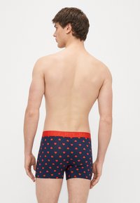 Tmavomodré boxerky s červenými vzormi srdiečok a červeným pásom s nápisom „HAPPY“ tučným písmom. Mäkká, pružná látka pre pohodlie.