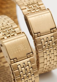 Goldfarbene Timex-Uhr mit gebürsteter Metalloberfläche, ausgestattet mit einem geflochtenen Armbanddesign und einer Schließe, detaillierter Kalender auf der Rückseite.