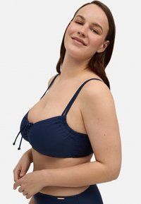 Top de bikini azul marino con tirantes ajustables, detalles fruncidos y acento de cuerda. Material suave y elástico con una textura lisa.