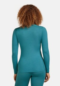 Osoba s kudrnatými vlasy, oblečená v těsně přiléhajícím tyrkysovém tričku s dlouhým rukávem a odpovídajících legínách, stojí zády k fotoaparátu.