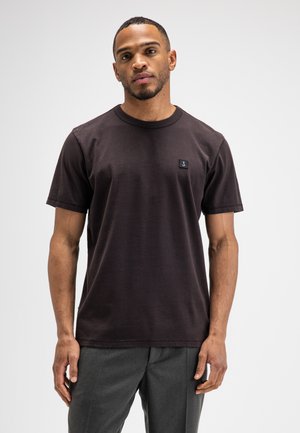 Mann steht, trägt ein einfarbiges schwarzes Kurzarm-T-Shirt mit kleinem Logo auf der Brust und dunkelgraue Hose vor einfachem Hintergrund.