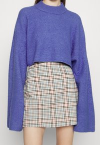 Pull court violet à manches larges, associé à une mini-jupe à carreaux aux couleurs pastel avec des accents marron. La texture douce en maille est visible.