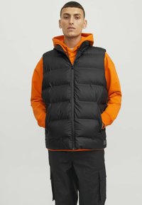 Sort puffer-vest med polstrede sektioner, lynlås foran og sidelommer, båret over en orange hættetrøje, parret med sorte cargo-bukser.