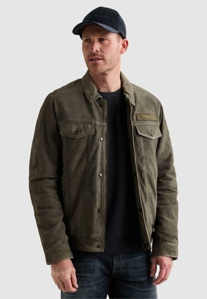 Homme portant une veste en daim vert foncé avec poches à rabat boutonnées, chemise noire, jean foncé et casquette de baseball noire regardant sur le côté.