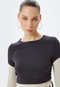 Zwart gebreid crop top met korte mouwen, voorzien van een zijkant met rimpeldetails en een contrasterend beige paneel aan de onderrand.
