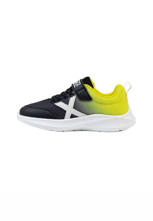 Zapatilla deportiva infantil negra y amarilla con suela blanca, diseño lateral en X blanco y cierre de velcro sobre fondo blanco.