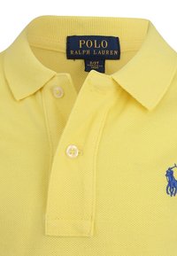 Gul polo skjorta med strukturerad tyg, två knappar vid kragen och en blå broderad logotyp på vänster sida. Varumärkesetikett synlig.