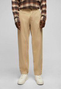 Beige Chinohosen mit einer glatten Textur, kombiniert mit weißen Sneakers. Das Hemd hat ein braun-graues Karomuster.