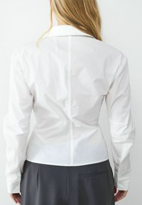Weißes, tailliertes Hemd mit spitzem Kragen, langen Ärmeln und geknöpften Manschetten. Maßgeschneiderter Schnitt mit glatter Textur und subtilen Nahtdetails auf der Rückseite.