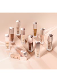 Un ensemble de dix bouteilles de fond de teint rectangulaires avec des bouchons métalliques rose doré. Les bouteilles affichent différentes nuances de fond de teint devant un fond clair.