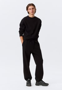 Jeune personne portant un sweat-shirt noir ample et un pantalon de survêtement assorti avec des baskets noires, debout les mains dans les poches devant un fond uni.