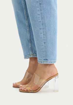 Füße in hellblauen Jeans und transparenten, hochhackigen Pantoletten mit klaren Blockabsätzen auf einem schlichten hellen Hintergrund.