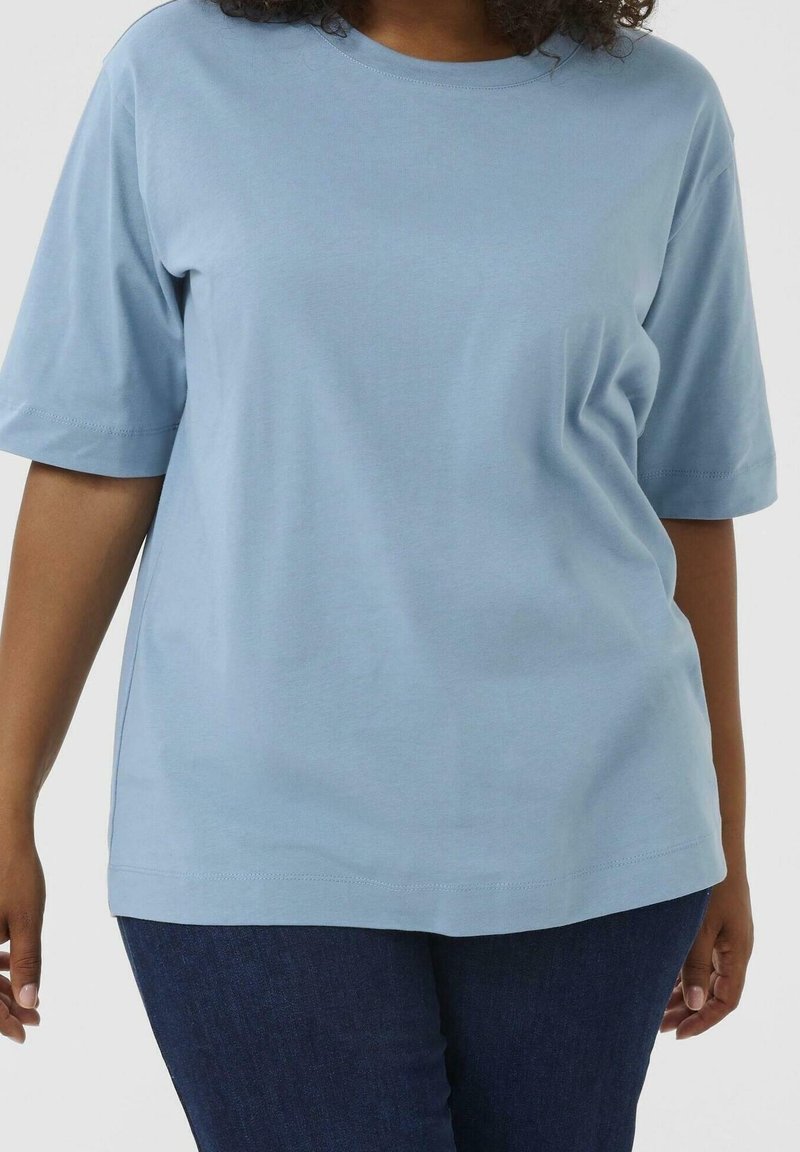 Femme portant un t-shirt bleu clair à manches courtes uni et un jean bleu foncé, debout devant un fond uni.
