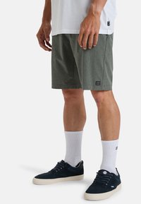 Pantaloni corti grigio-verdi con una texture sottile, dotati di una toppa con logo, abbinati a calze bianche e sneakers blu scuro con suole chiare.