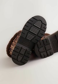 Brązowe botki z futra syntetycznego w leopardzi wzór z czarną gumową podeszwą, z teksturowanymi podeszwami dla lepszej przyczepności i charakterystycznym logo "NEXT" na podeszwie.