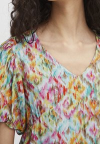 Blouse colorée avec un col en V, présentant un motif de rose, jaune, bleu et marron. Le tissu a une apparence douce et légèrement texturée.