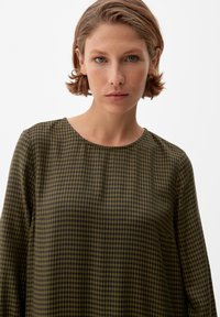 Dunkelgrüne und schwarze karierte Bluse mit Rundhalsausschnitt und langen Ärmeln, hergestellt aus einem glatten, strukturierten Stoff.