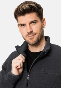 Veste en polaire gris foncé avec un col montant et fermeture à demi-zip. Matériau texturé et détails de couture proéminents. Logo de la marque visible sur la poitrine.