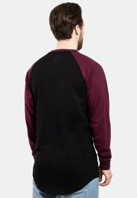 Majica s dugim rukavima koja ima crno tijelo s burgundskim raglan rukavima. Mekani materijal, zaobljeni rub i suptilan detalj s logom na stražnjoj strani.