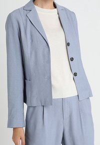 Jasnoniebieska, dopasowana marynarka z wyciętymi klapami, czterema guzikami z przodu i bocznymi kieszeniami. Nosi się ją na białym swetrze, tkanina wydaje się mieć teksturę.