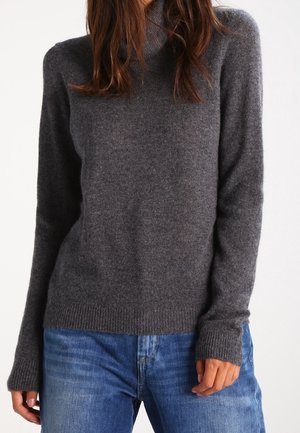 Kvinde iført en mørkegrå rullekravebluse med lange ærmer og blå jeans, stående mod en ensartet hvid baggrund.