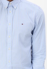 Camicia a quadri blu chiaro con colletto button-down, bottoni bianchi e una piccola etichetta con logo. Il tessuto appare liscio e fresco.
