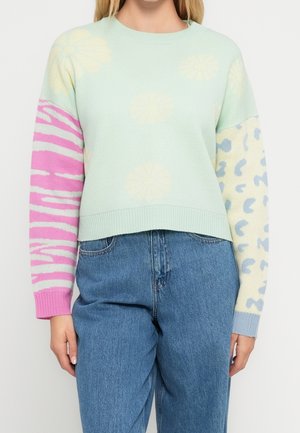 Kvinde iført en pastelgrøn trøje med cremefarvede blomster, lyserød zebra print på venstre ærme, blå og cremefarvet leopard print på højre ærme og blå jeans.