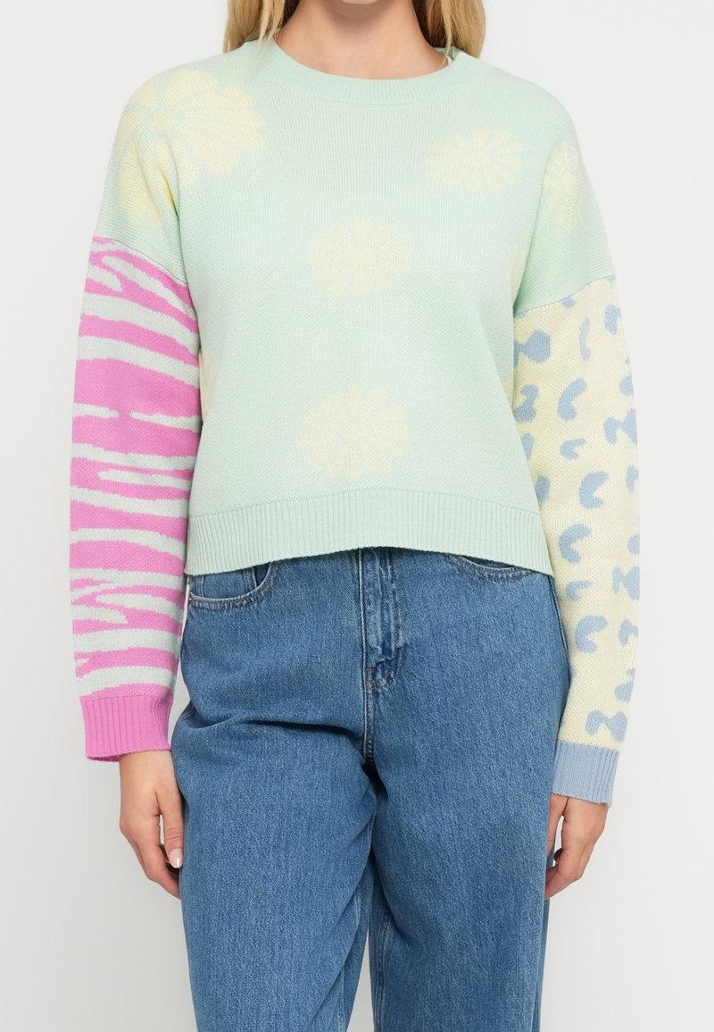 Femme portant un pull vert pastel avec des fleurs crème, manche gauche imprimé zèbre rose, manche droite imprimé léopard bleu et crème, et jean bleu.