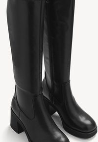 Bottes noires montantes en cuir lisse. Elles sont dotées d'un talon épais, d'une fermeture zippée sur le côté et de panneaux élastiques pour plus de confort.