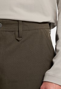 Pantalons verts foncés texturés avec un devant plat, des passants de ceinture et un motif à carreaux subtil, associés à un haut à manches longues de couleur claire.