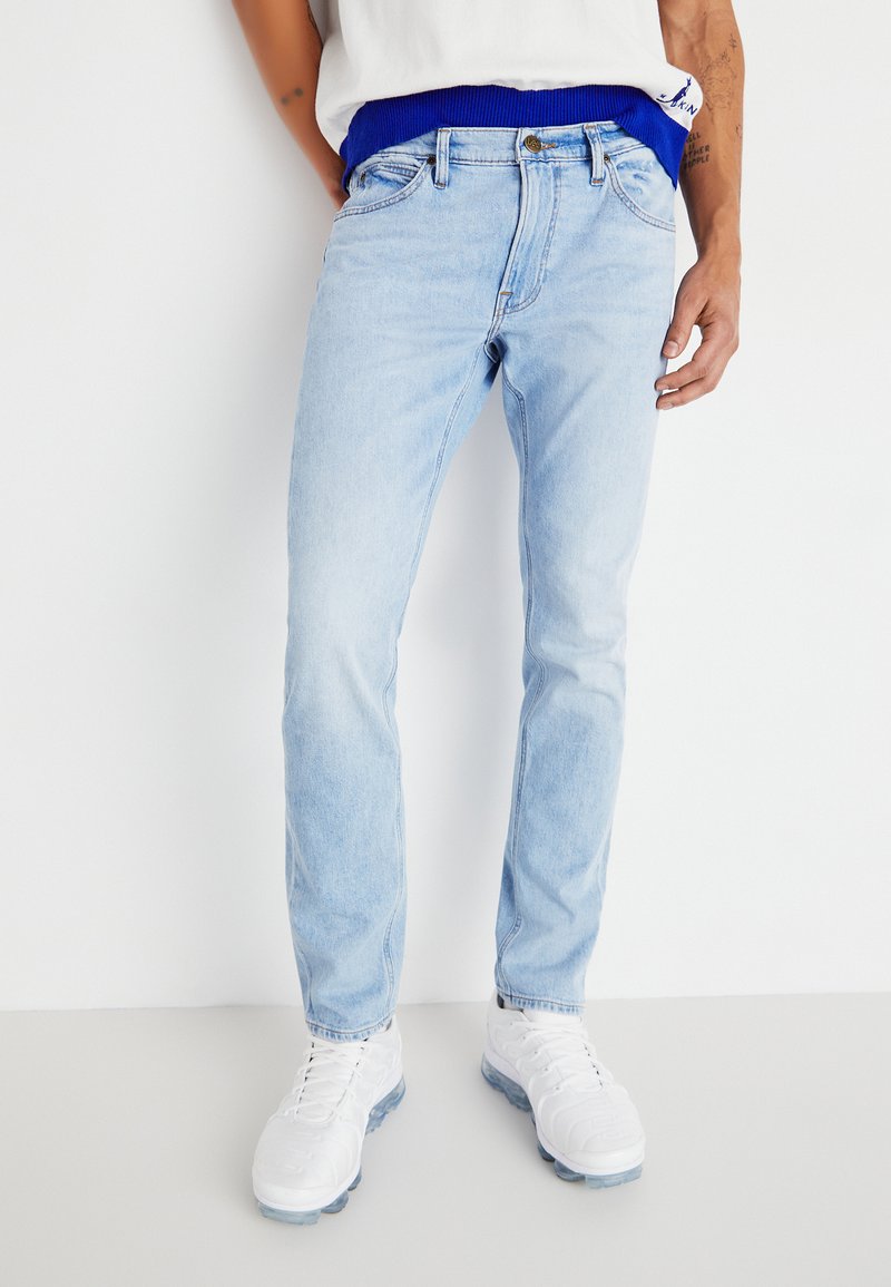 Los jeans de mezclilla azul claro presentan un diseño clásico de pierna recta con un ligero desgaste, acentuados por un botón plateado y cierre de cremallera.